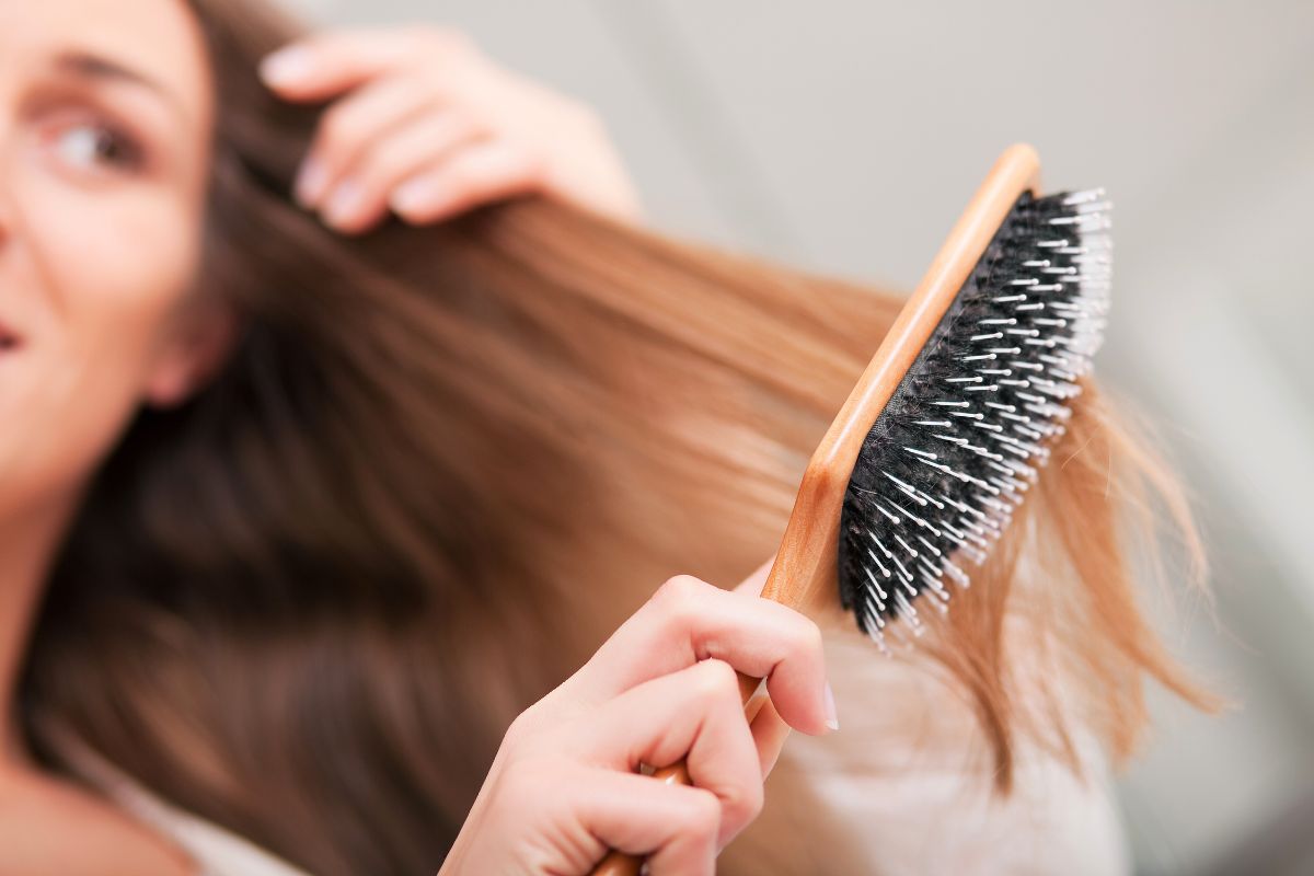 Does brushing hair stimulate growth: woman brushing long healthy brown hair with wooden paddle brush to distribute natural oils and promote scalp health