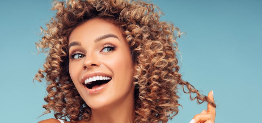 Joyful young woman with bright eyes and radiant smile holding a loose curl of her voluminous, bouncy noodle hair against a fresh blue background, showcasing perfect curly texture.