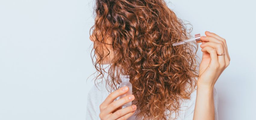 Woman with long, shiny brown noodle hair applying a clear hair serum with a dropper, treating her defined curls for extra hydration and bounce in soft natural lighting.