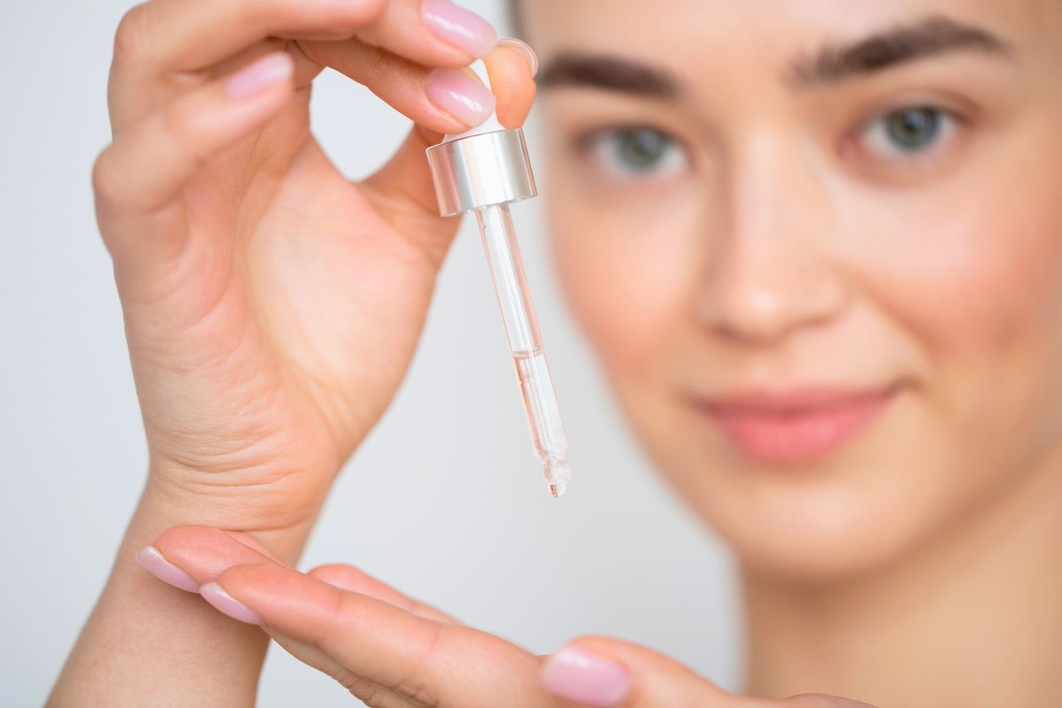 Woman examining a drop of clear lash serum from a dropper bottle on her fingertip, illustrating precise application of lash serums for eyelash growth and conditioning