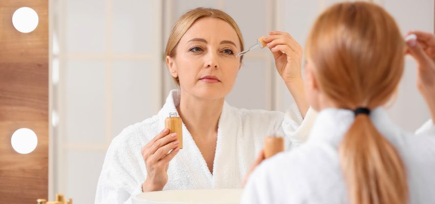 Woman in robe carefully applying lash serum to her upper lashes with a precision wand in front of a vanity mirror, demonstrating daily use of lash serums for fuller appearance